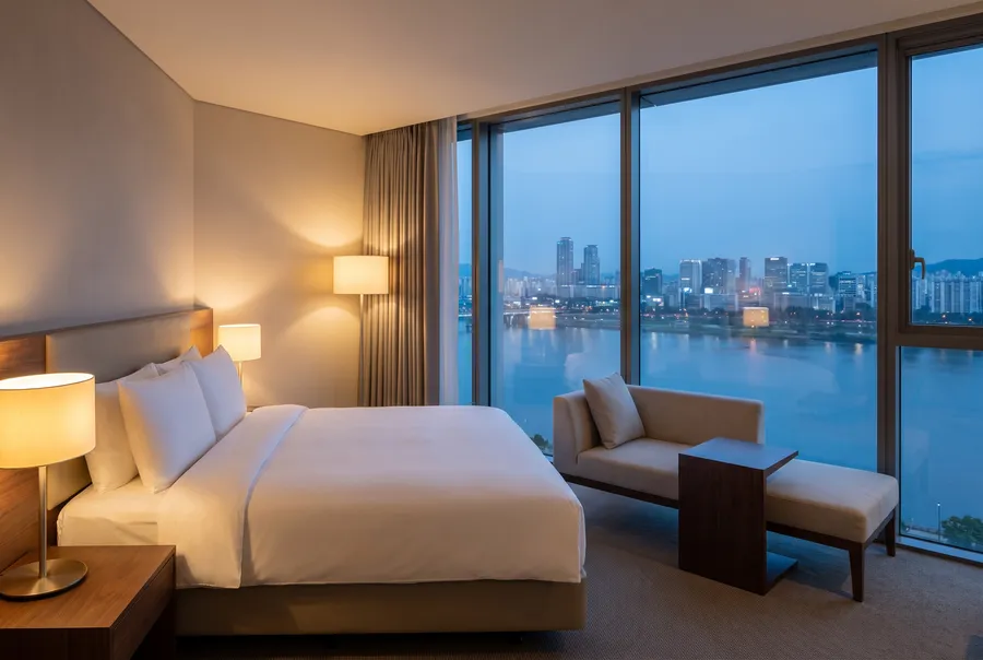 Generated scene: a tidy hotel room at dusk with a white bed, lamps and a chaise by a floor-to-ceiling window over a river and a city skyline.